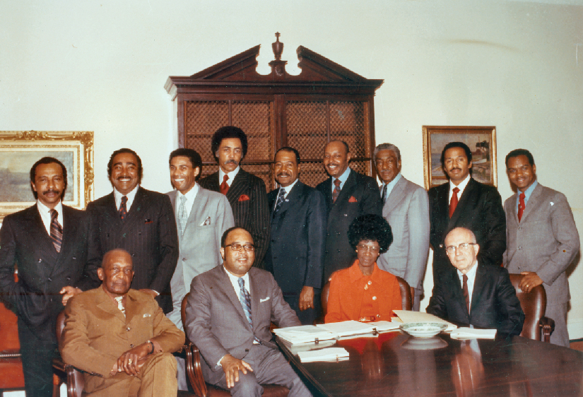 An image of a group of people, four of whom are seated at a table, and nine of whom are standing.