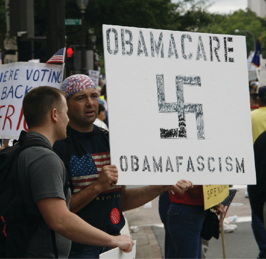 An image of a person holding a sign that reads “Obamacare obamafascism” and has the symbol of a swastika.