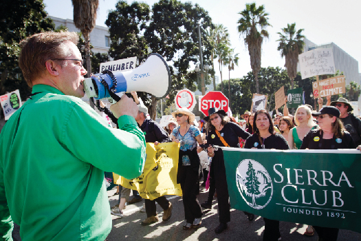 An image of a person speaking through a bullhorn on the left, and a crowd of people marching down a street on the right. Several marchers are holding a large banner that reads “Sierra Club”.