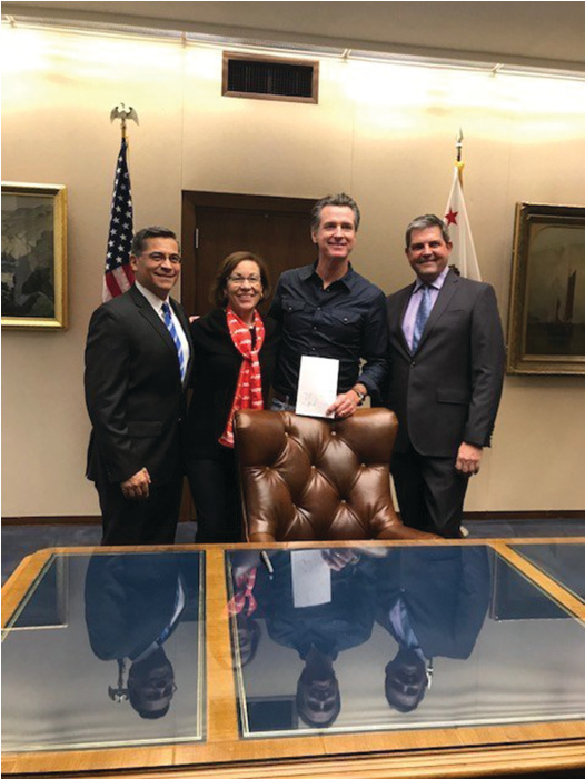 Four people stand behind a desk: (Left to Right) Attorney General Becerra, AARP Advocacy Manager Blanca Castro, Governor Newsom, and Assemblymember Jim Wood. Newsom holds a document.