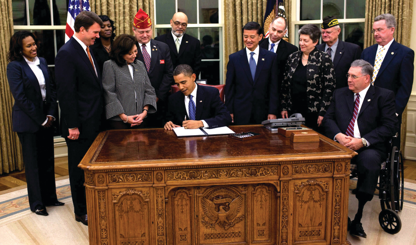 An image of a group of 12 people standing around Barack Obama, who is seated at a desk and signing a piece of paper.