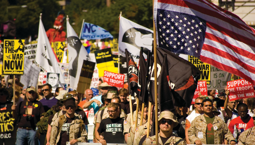 An image of a group of people, several of whom are holding flags and signs. One of the signs reads “End the war now”, and another reads “Support the troops, end the war”.