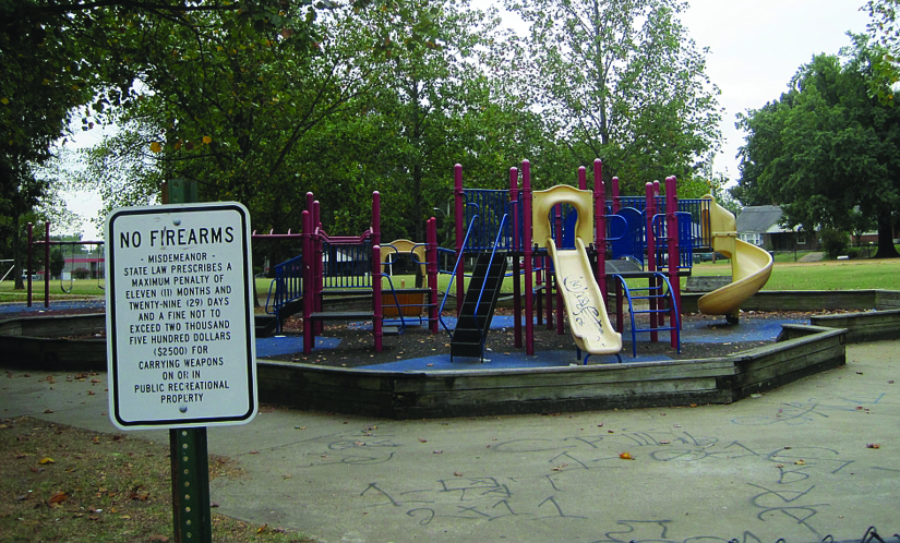 A photo of a sign that reads “No Firearms”. A playground can be seen in the background.
