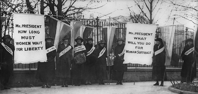 An image of several people standing in front of a fence. Some people are holding banners. The banners read “Mr. President how long must women wait for liberty” and “Mr. President what will you do for woman sufferage”.
