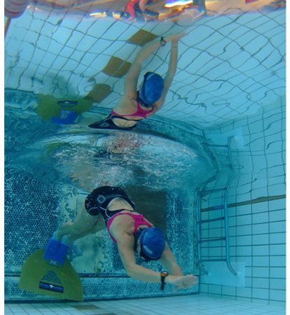 The figure is a photograph taken from underwater. The photo shows an underwater swimmer. Above the swimmer is an upside down image of the swimmer and of the activities on the deck, outside the pool.