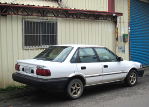 Old white Geo brand car parked outside of a building.