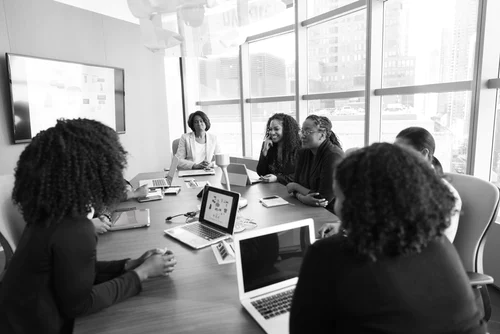 Black and white image of six smiling women sit around a table in a conference room with laptops.