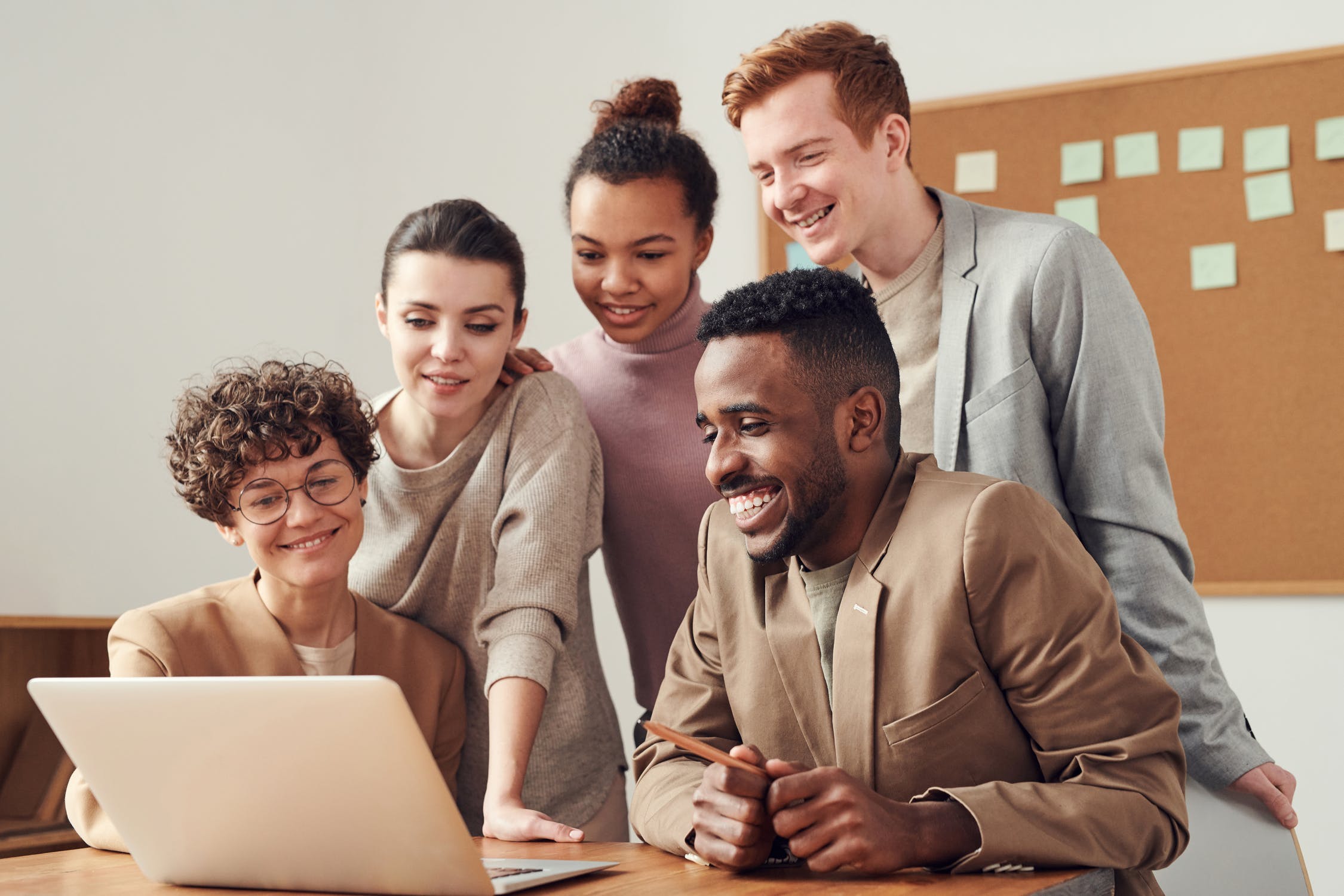 Five young adults looking at a laptop with a bulletin board in the background.