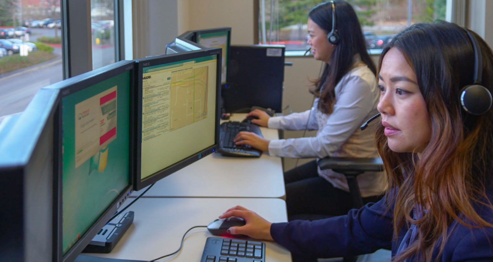 Two females sit at a desk in front of computers with head sets on.