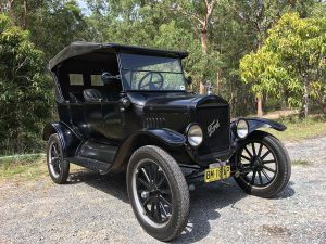 Black 1925 Ford Model T car with a yellow license plate parked outside near trees.