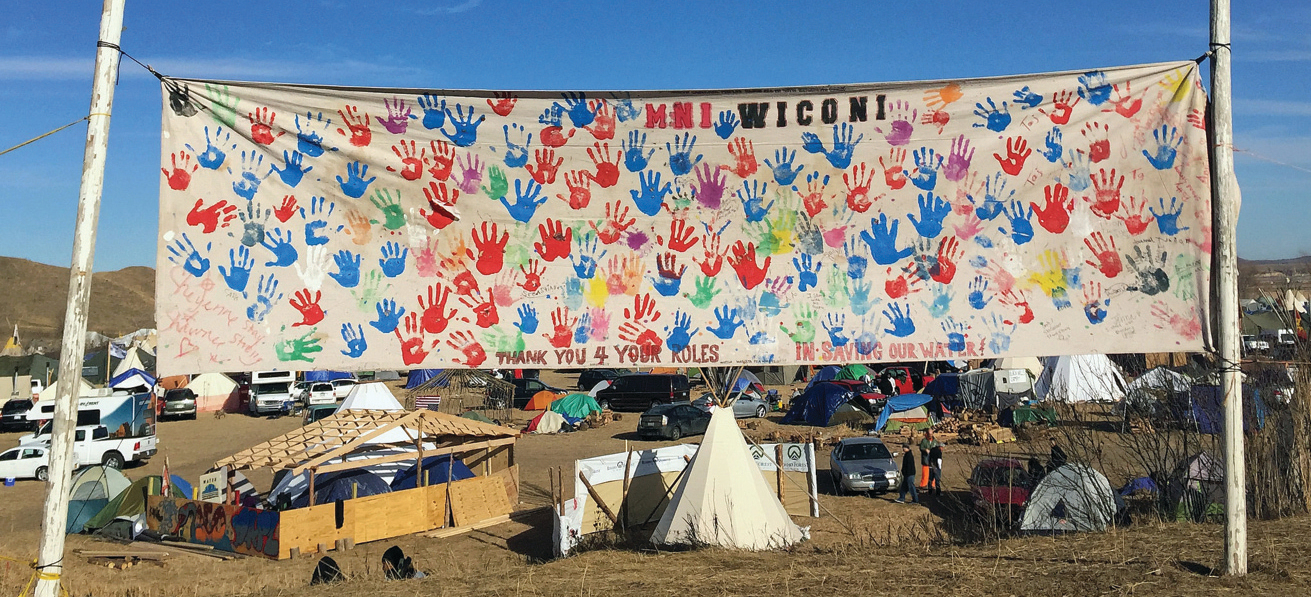 A camp settlement shows several vehicles, tents, and a teepee. A large banner covered with brightly colored handprints reads 'MNI WICONI' and 'Thank You 4 Your Roles in Saving Our Water'.