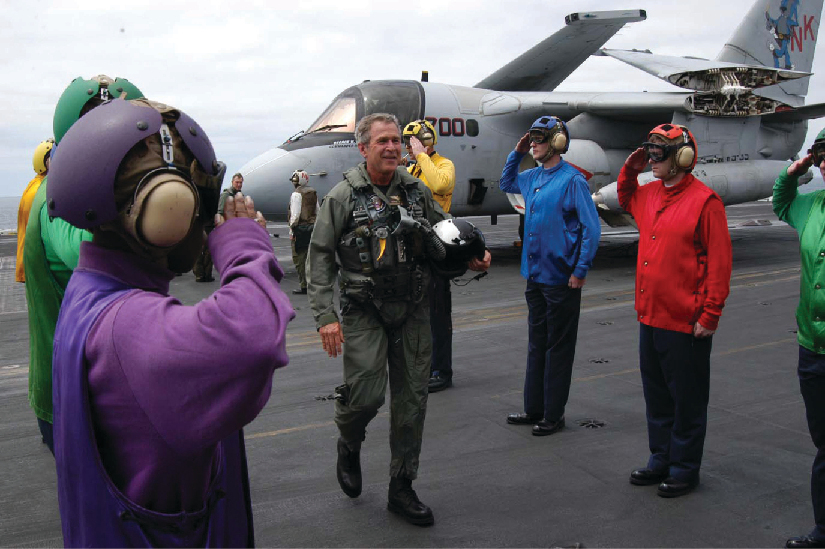 A photo of George W. Bush in a flight suit stepping out of a plane onto an aircraft carrier. Personnel stand on either side and salute him.