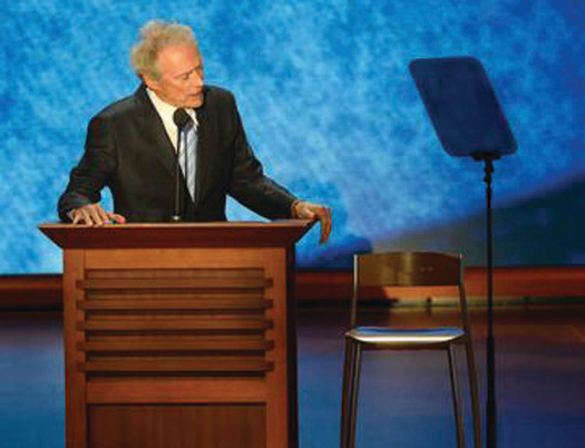 An image of Clint Eastwood standing behind a podium. Next to him on the right is an empty chair.