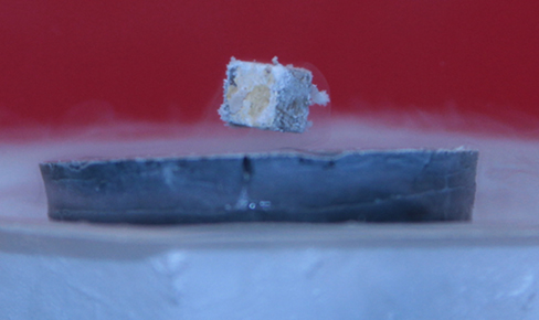 Picture shows a photograph of a small magnet in a shape of cube levitating over the surface of a superconductor.