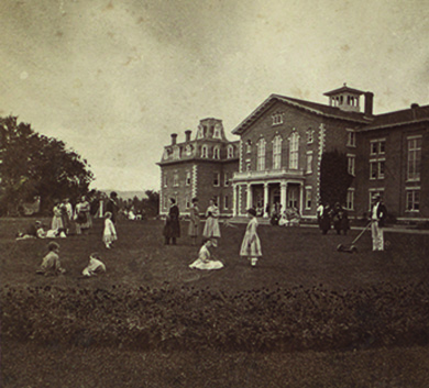A photograph depicts a large, opulent house surrounded by a lawn on which men, women, and children sit, stand, and converse.