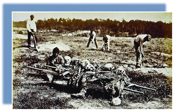 A photograph shows several African American men collecting the bones from a battleground in Virginia. In the foreground, one man prepares to carry away a pile of skulls and body parts.