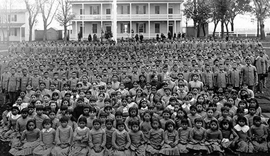 A photograph shows a large, posed group of Native American children at an Indian school. The girls sit in the front in collared dresses. The boys stand at the back in button-down shirts and slacks.