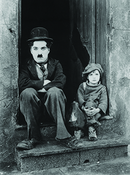 Charlie Chaplin is shown sitting in a doorway with his arms folded, accompanied by a small, shabbily dressed child.