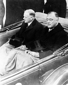 A photograph shows Herbert Hoover and Franklin D. Roosevelt riding side-by-side in the back of a convertible vehicle. A blanket covers their legs, and their hats rest on their laps.