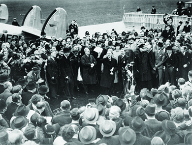 A photograph shows Neville Chamberlain immediately following his arrival in England, where he addresses an enthusiastic crowd of officials and press.