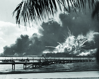 A photograph shows a long dock with the USS Shaw exploding behind it. In the far background, massive billows of smoke are visible.