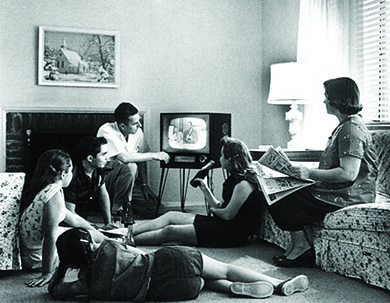 A photograph shows a man, a woman, three teenage girls, and a teenage boy sitting in a living room, watching a television.