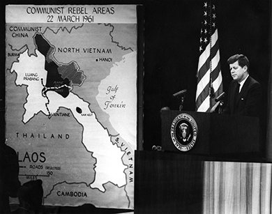 A photograph shows President Kennedy standing at a podium delivering a speech. Beside him hangs a large map of Southeast Asia, labeled “Communist Rebel Areas/22 March 1961.”