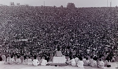 A photograph depicts the rear view of a stage, in front of which a massive crowd is gathered. Swami Satchidananda sits cross-legged atop a podium, with a small group of others seated on the stage to each side of him.