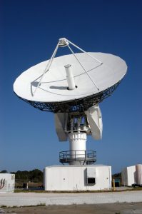 The large, round dish antenna looking like a giant white saucer is shown. It rests on a pillar shaped structure with a moveable tracking system that allows it to point towards a target object, send out electromagnetic waves, and collect any signals that bounce back from the target object.