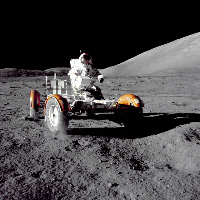 Photograph of the lunar rover on the Moon. The photo looks like it was taken at night with a powerful spotlight shining on the rover from the left: light reflects off the rover, the astronaut, and the Moon’s surface, but the sky is black. The shadow of the rover is very sharp.