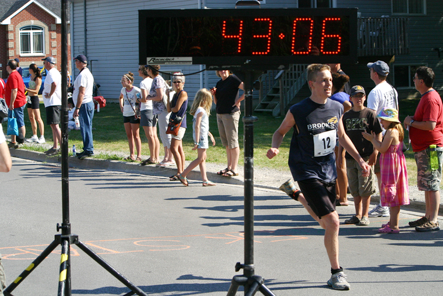 A runner crossing a finishing line on a road with a clock showing his finish time.