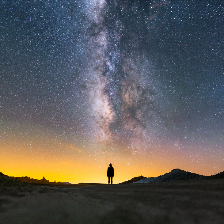Image of the night sky showing the Milky Way, a dense, vertical band of stars. Under the Milky Way is the silhouette of a person.