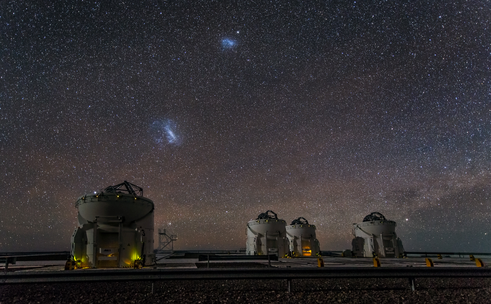 The Magellanic Clouds. The Large Magellanic Cloud is to the left of center and the SMC just above center in this image taken at Cerro Paranal in Chile. Four of the VLT’s auxiliary telescopes are seen in the foreground.